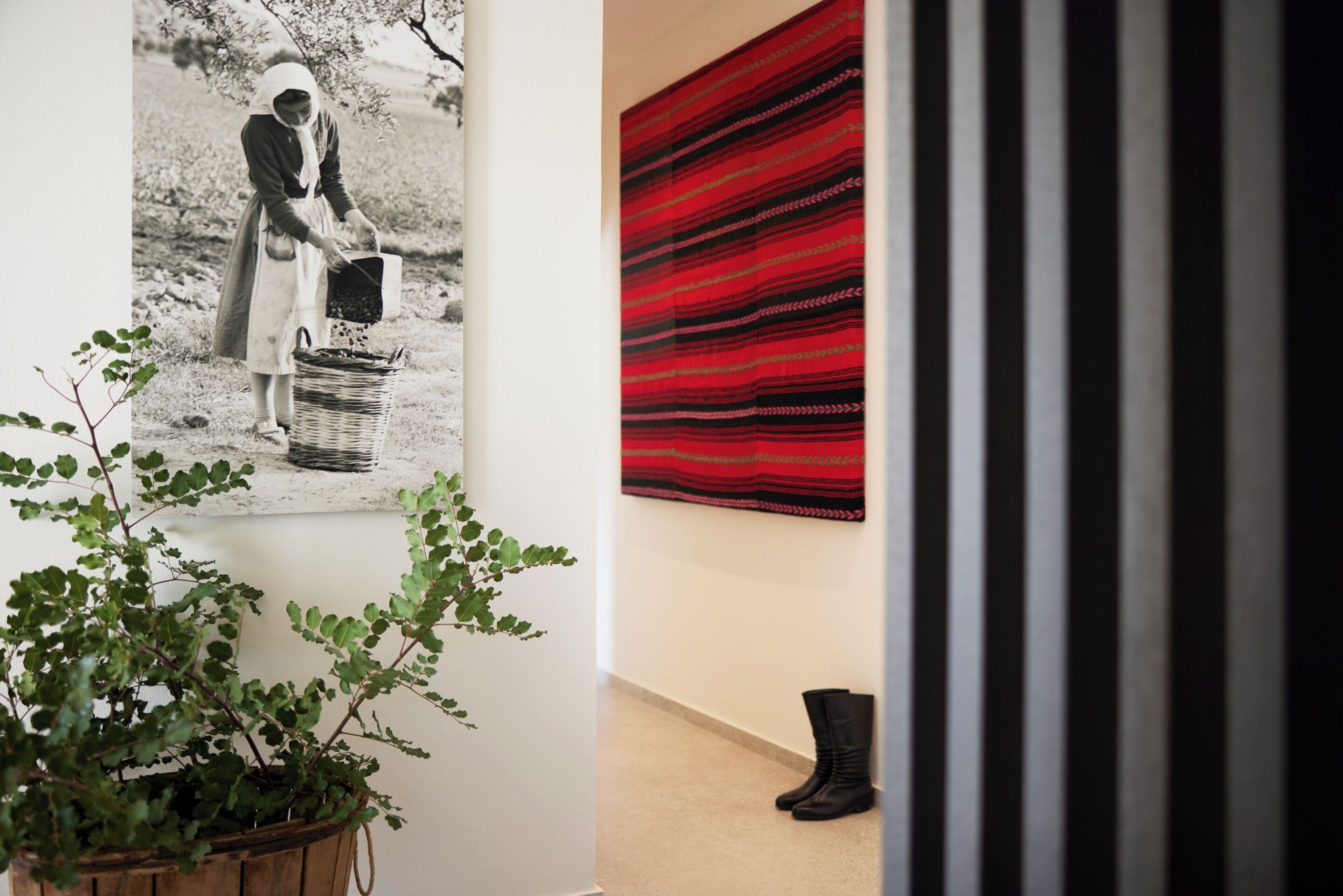 Une pièce dans laquelle on trouve une photographie accrochée au mur, une plante en pot, un tissu rouge à motifs et une paire de bottes.