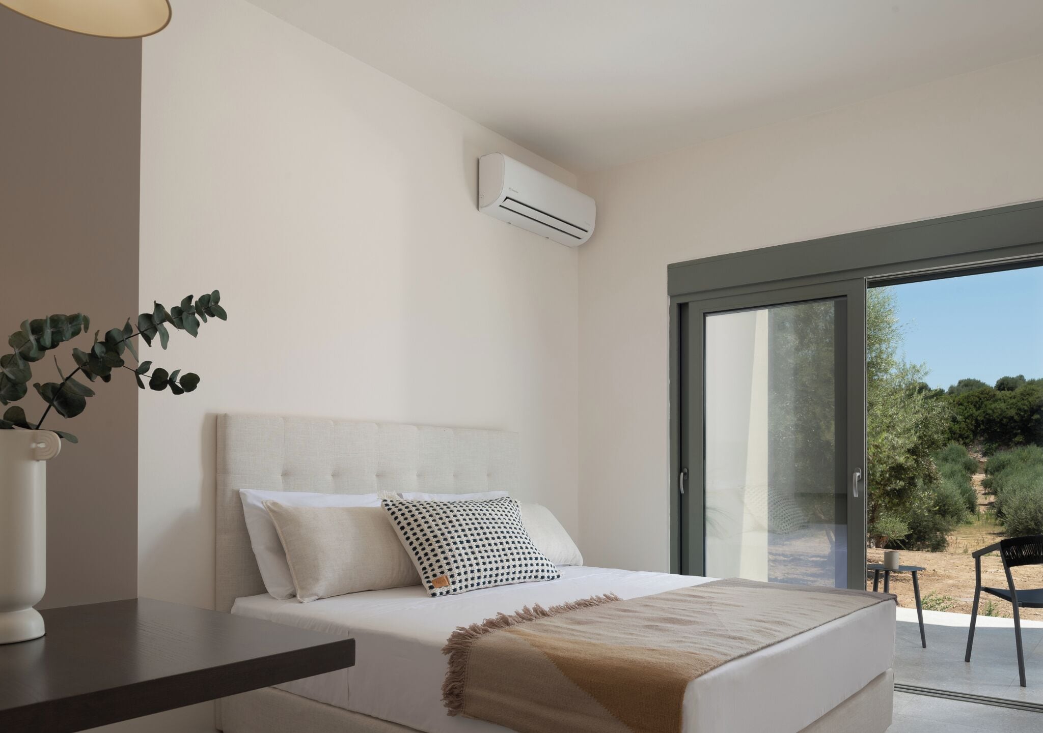 Een slaapkamer met een bed, kussens, een nachtkastje, een vaas met een plant, een airconditioning en uitzicht door een glazen deur.
