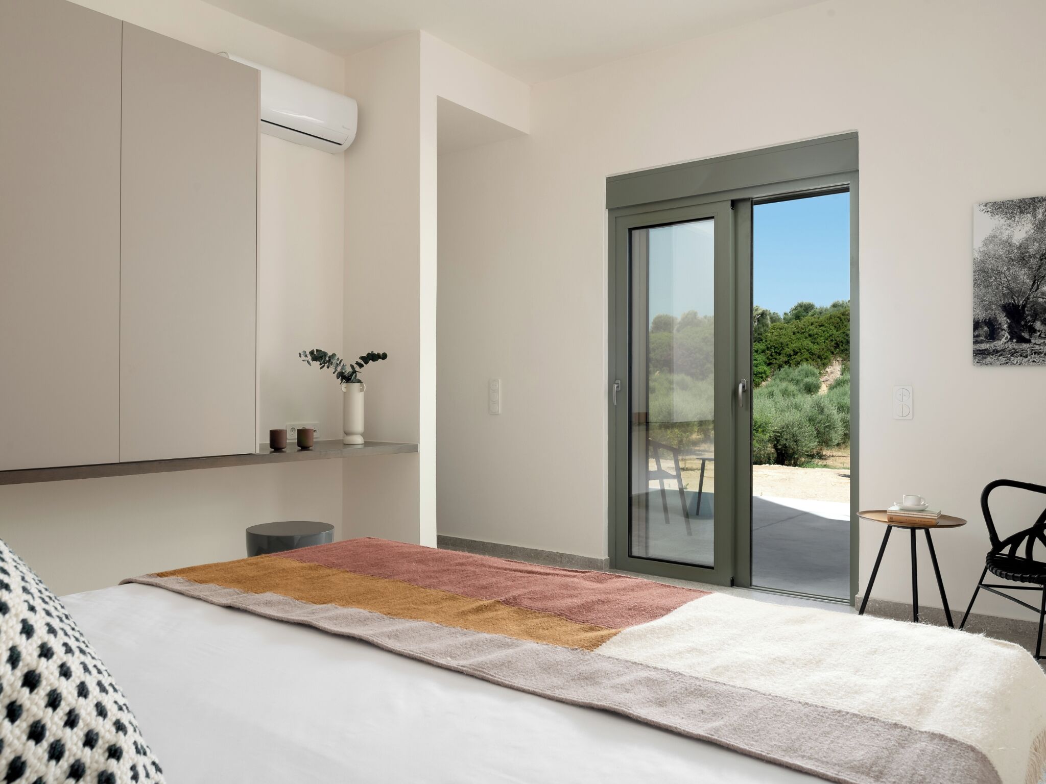 A bedroom with a bed, a nightstand, a window with a view of greenery, and a sliding glass door leading outside.