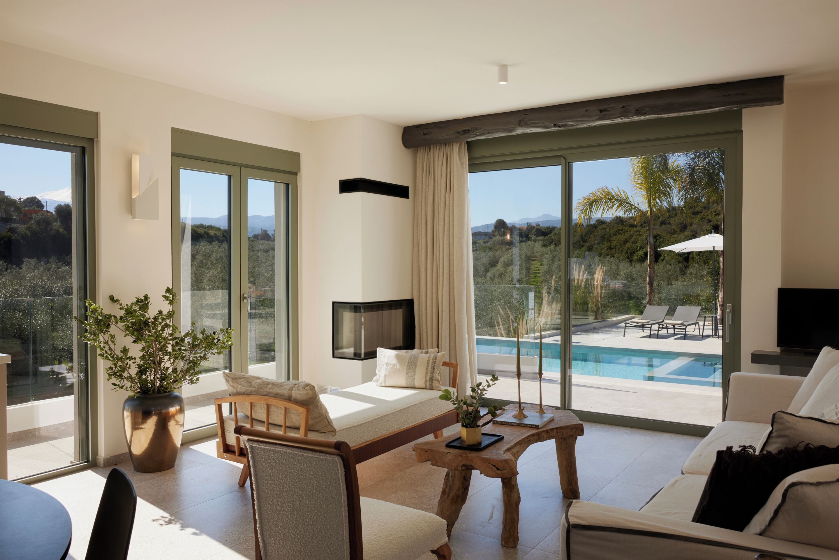 A modern living room with a sofa, a coffee table, a TV, and a large window overlooking a pool and landscape.