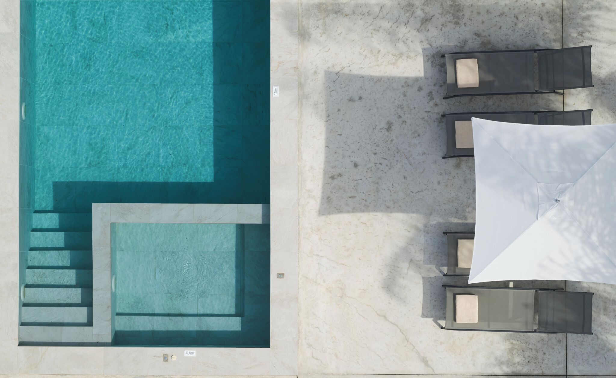 An aerial view of a swimming pool with steps leading into it, a sun lounger area with umbrellas, and a tiled patio.
