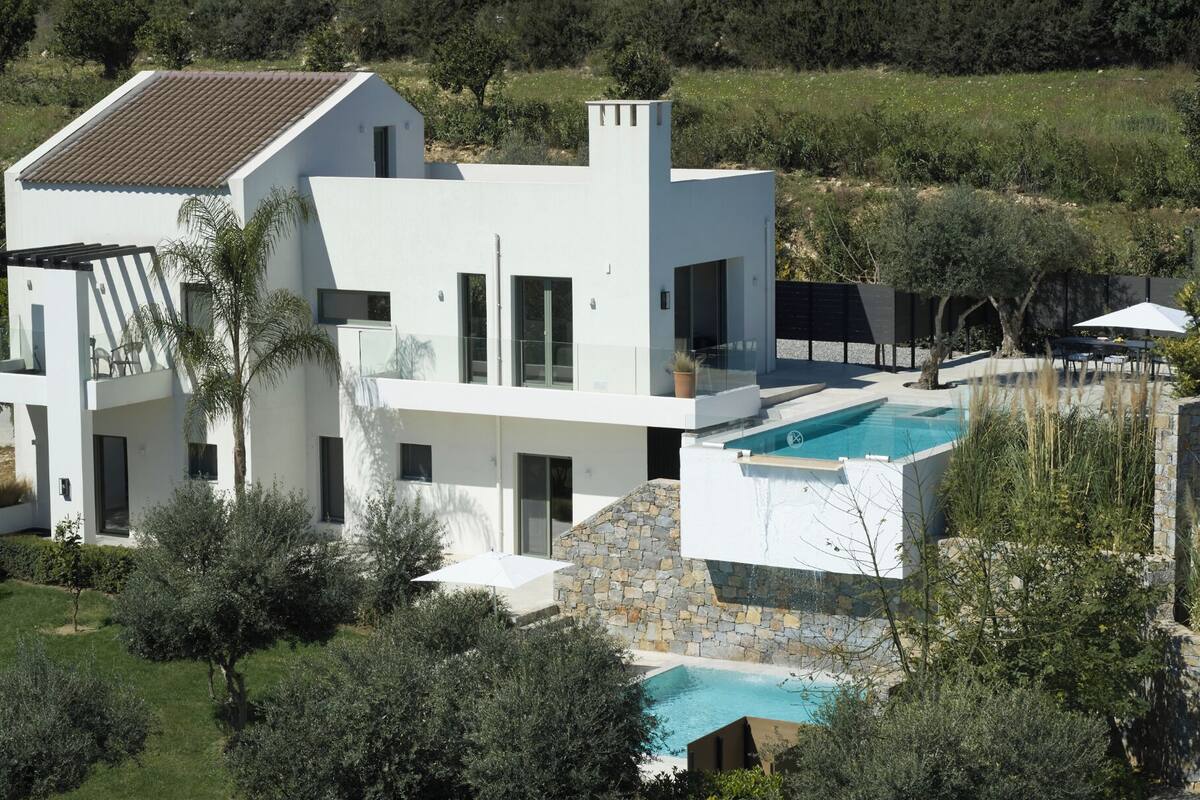 A modern two-story house with a swimming pool, surrounded by greenery and a stone wall.