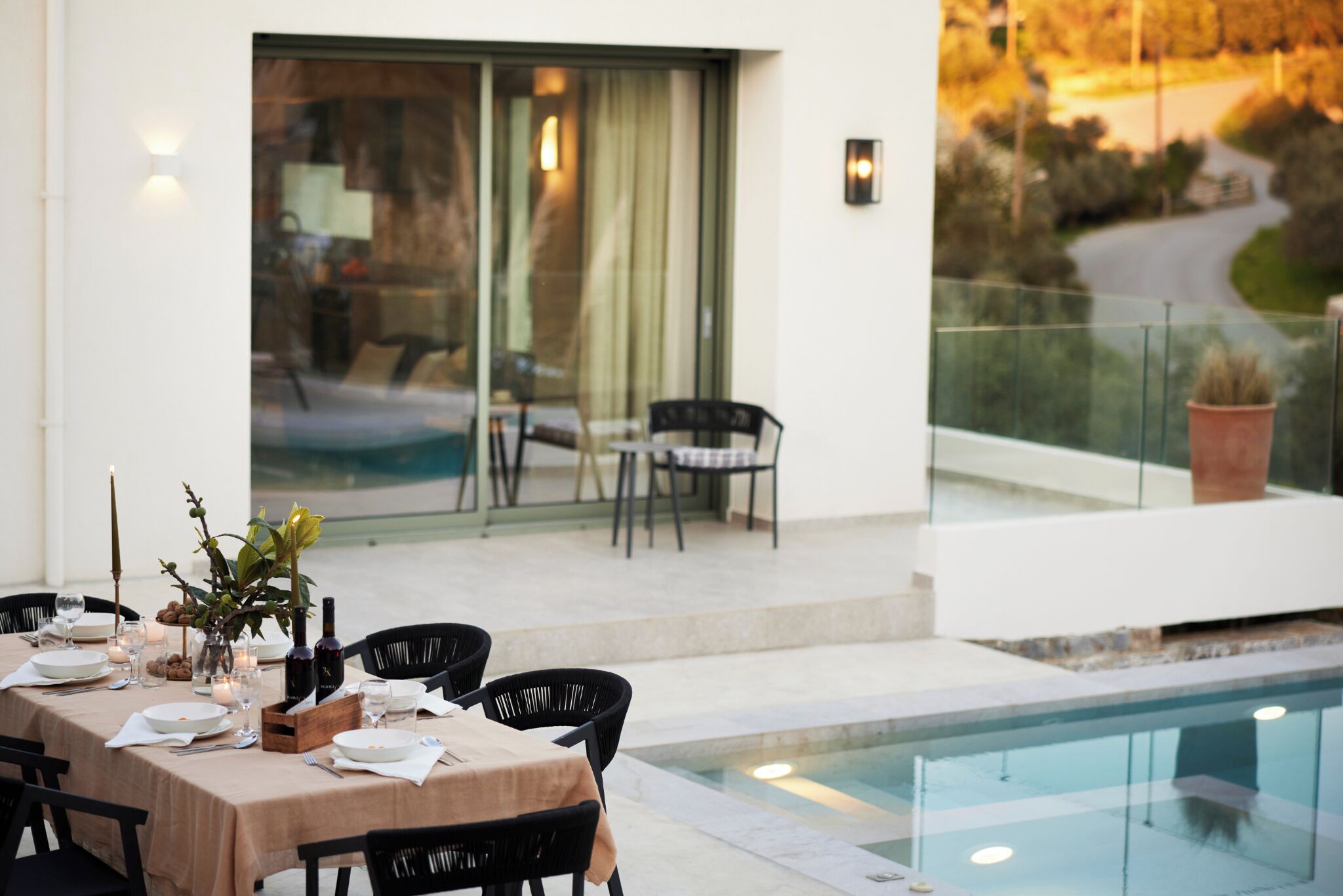 Un coin repas avec une table dressée pour quatre personnes, des chaises, une piscine et une vue sur une route et la verdure.