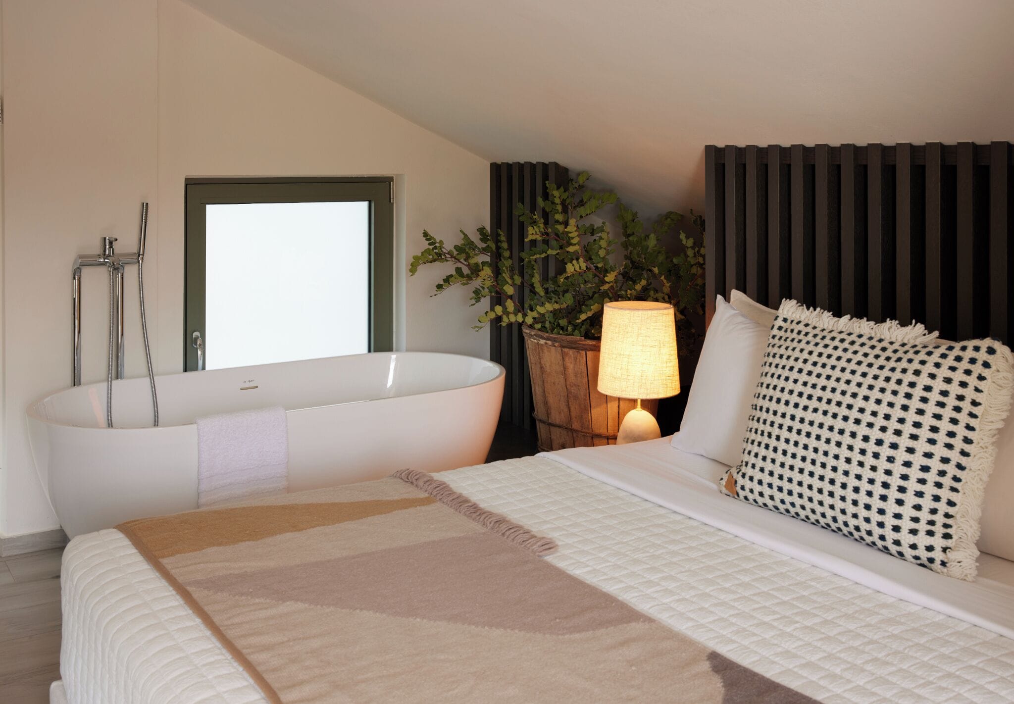 A modern hotel room with a large bathtub, a bed with a beige and white quilted bedspread, a bedside lamp, and a potted plant.
