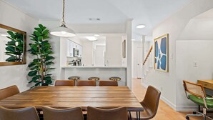 A dining area with a wooden table, brown chairs, a hanging plant, and a kitchen in the background. - Fly Creek Collection - Sunset Perch (Fairhope)