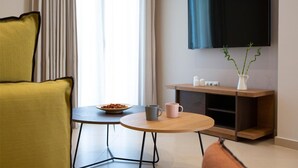 Una habitación de hotel moderna con un sillón amarillo, una pequeña mesa redonda con un tazón de cereal y dos tazas, un mueble bajo de madera con un televisor encima y una ventana con cortinas.