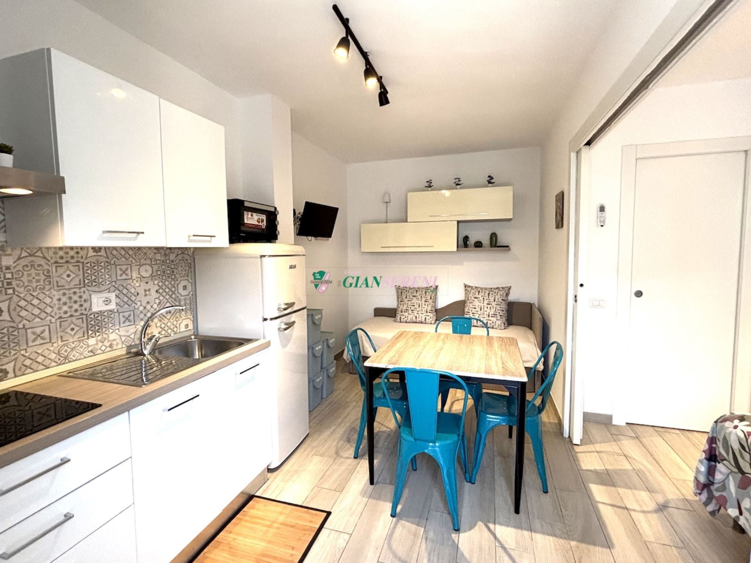 Une cuisine compacte avec des meubles blancs, une cuisinière encastrée et un coin repas comprenant une table en bois et des chaises bleues.