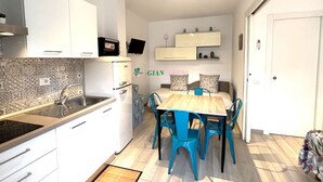 Cocina compacta con armarios blancos, estufa integrada y comedor con mesa de madera y sillas azules.
