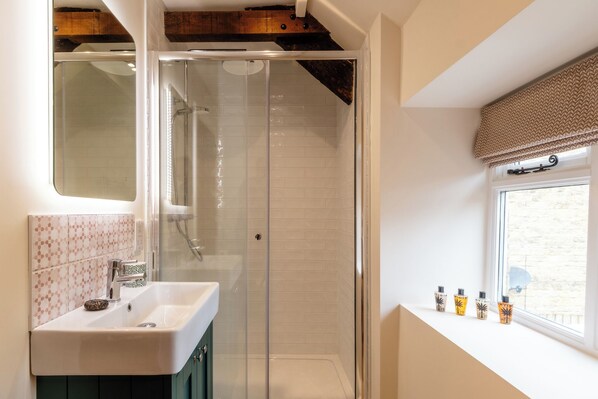 A bathroom with a glass shower enclosure, a sink with a decorative soap dish, and a window with a view. - The Cowshed Banham Cottages (Bourton-on-the-Water)
