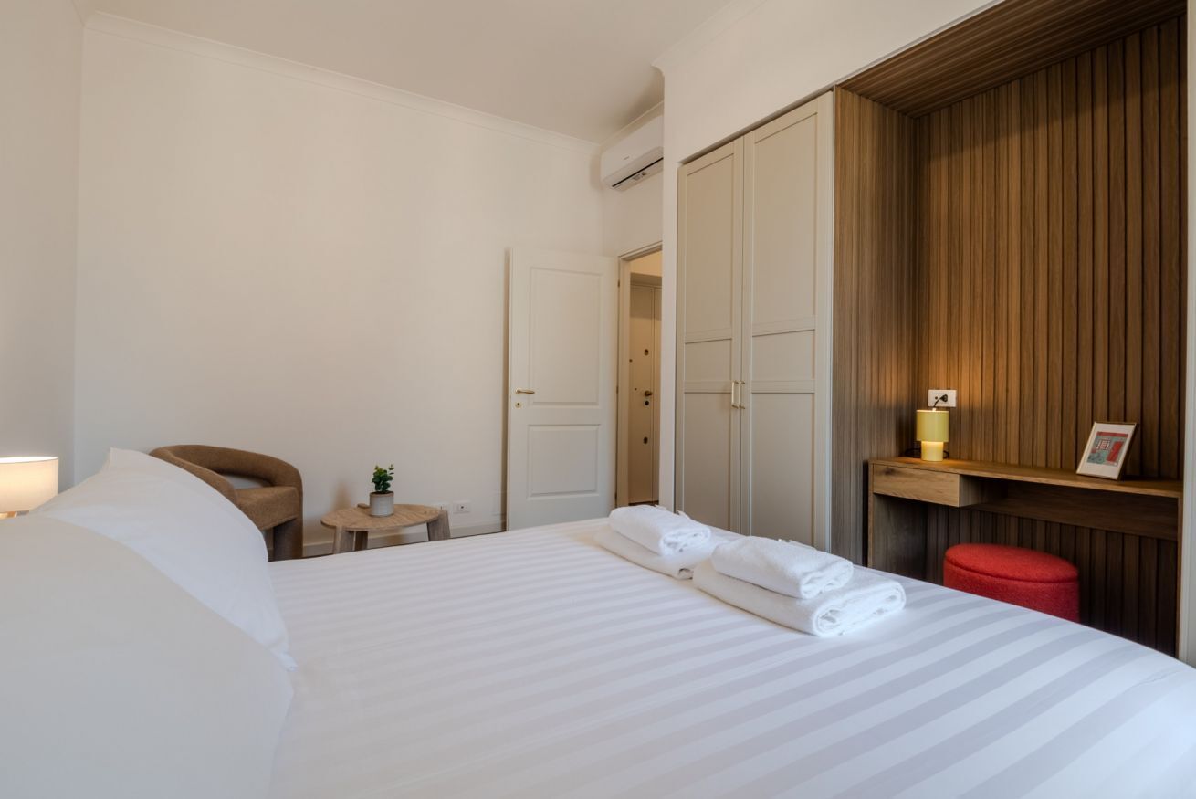 Quarto de hotel com cama, uma cadeira, uma mesinha com uma planta, um banquinho vermelho e um guarda-roupa de madeira.