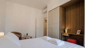 Une chambre d’hôtel avec un lit, une chaise, une petite table avec une plante, un pouf rouge et une armoire en bois.