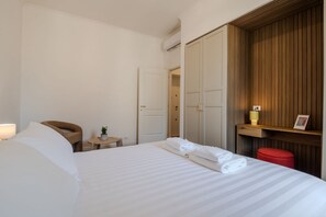 Ein Hotelzimmer mit einem Bett, einem Sessel, einem kleinen Tisch mit einer Pflanze, einem roten Hocker und einem Holzschrank.