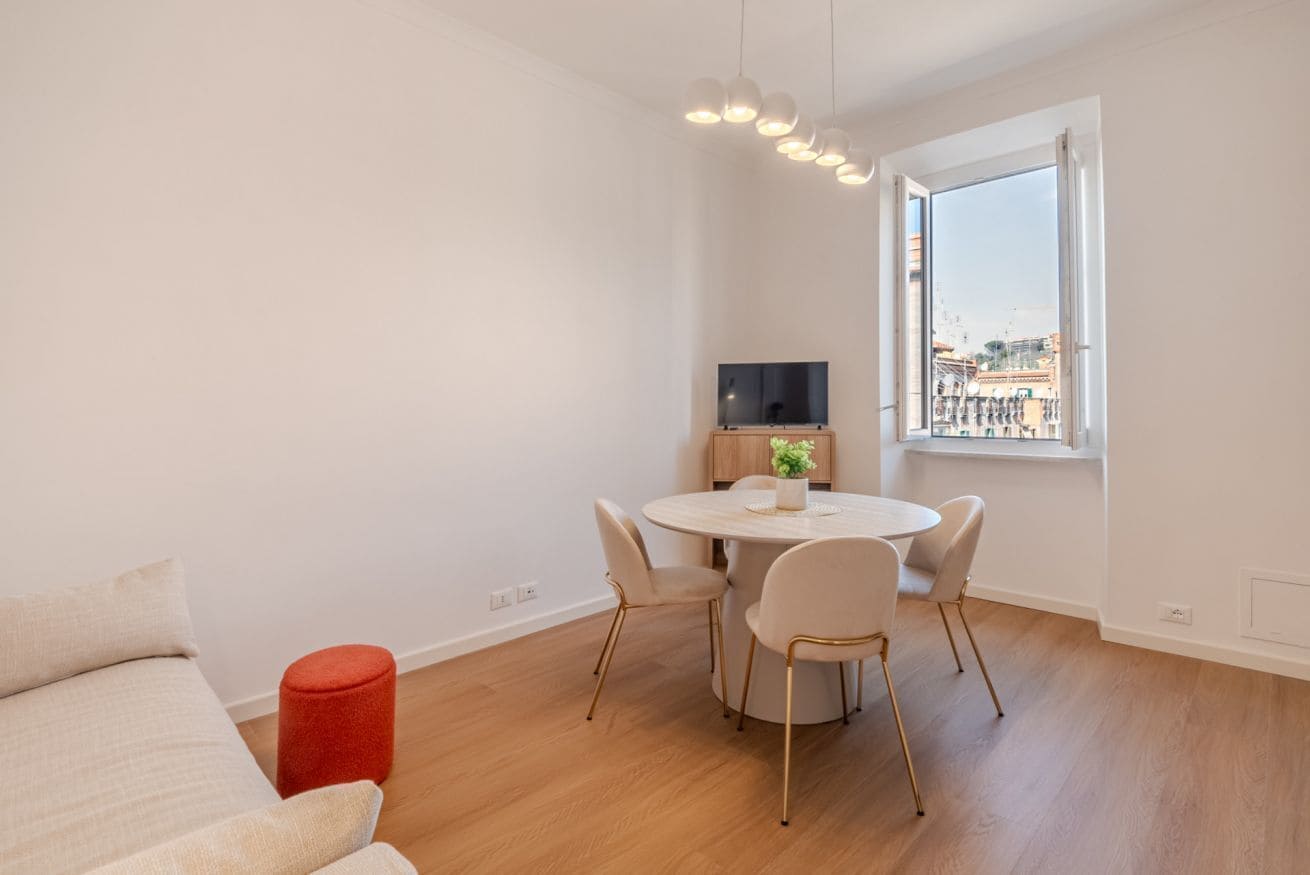 Una sala moderna con una mesa redonda de comedor, un sofá, un reposapiés rojo, un televisor y una ventana con vista a la ciudad.