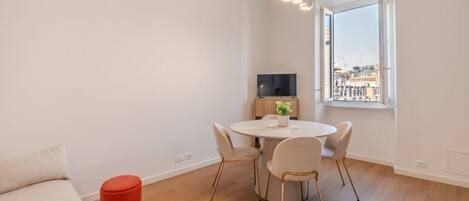 Ein modernes Wohnzimmer mit einem runden Esstisch, einer Couch, einem roten Hocker, einem Fernseher und einem Fenster mit Blick auf die Stadt.