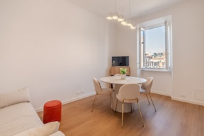 Ein modernes Wohnzimmer mit einem runden Esstisch, einer Couch, einem roten Hocker, einem Fernseher und einem Fenster mit Blick auf die Stadt.
