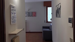 A hallway with a wooden floor, a grey sofa, and a radiator. There are wall-mounted lights and a thermostat.