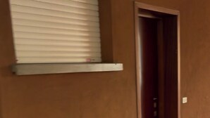 A hallway with a door, a window with blinds, and a wall-mounted shelf.