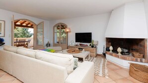 Een woonkamer met een bank, een televisie, een open haard en een eethoek met een tafel en stoelen.