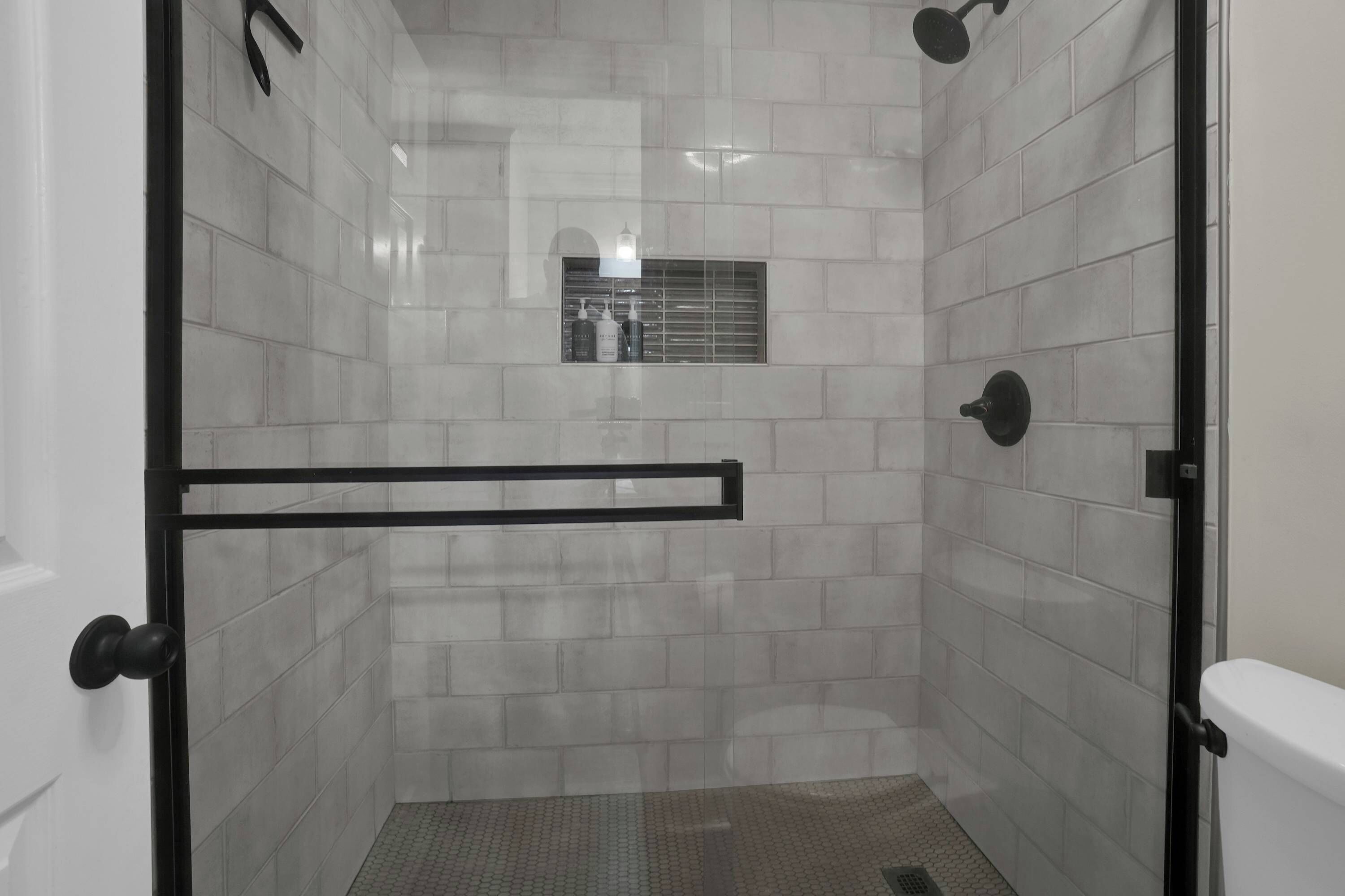 Un box de ducha con paredes de azulejos blancos, una manija de puerta negra y un pequeño ventanal con botellas.
