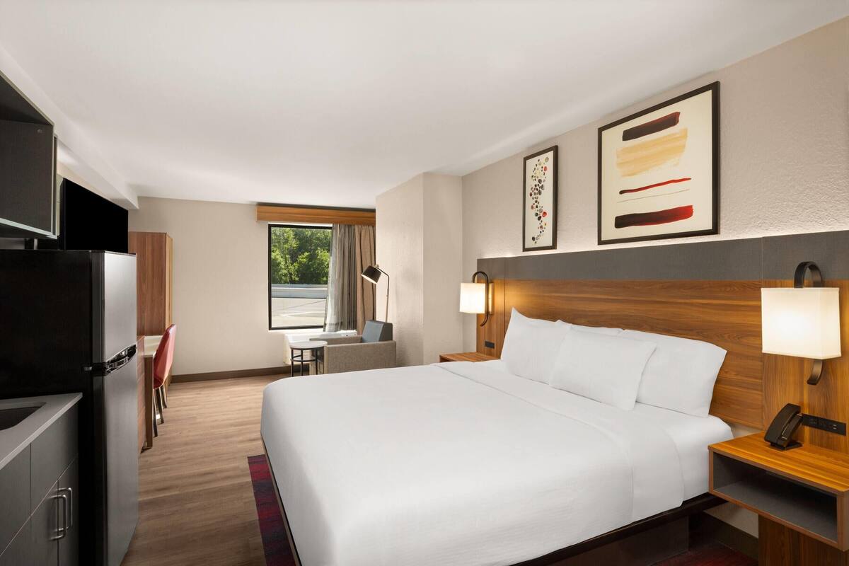 Quarto de hotel com cama, mesa de cabeceira, abajur, televisão e escrivaninha com cadeira.