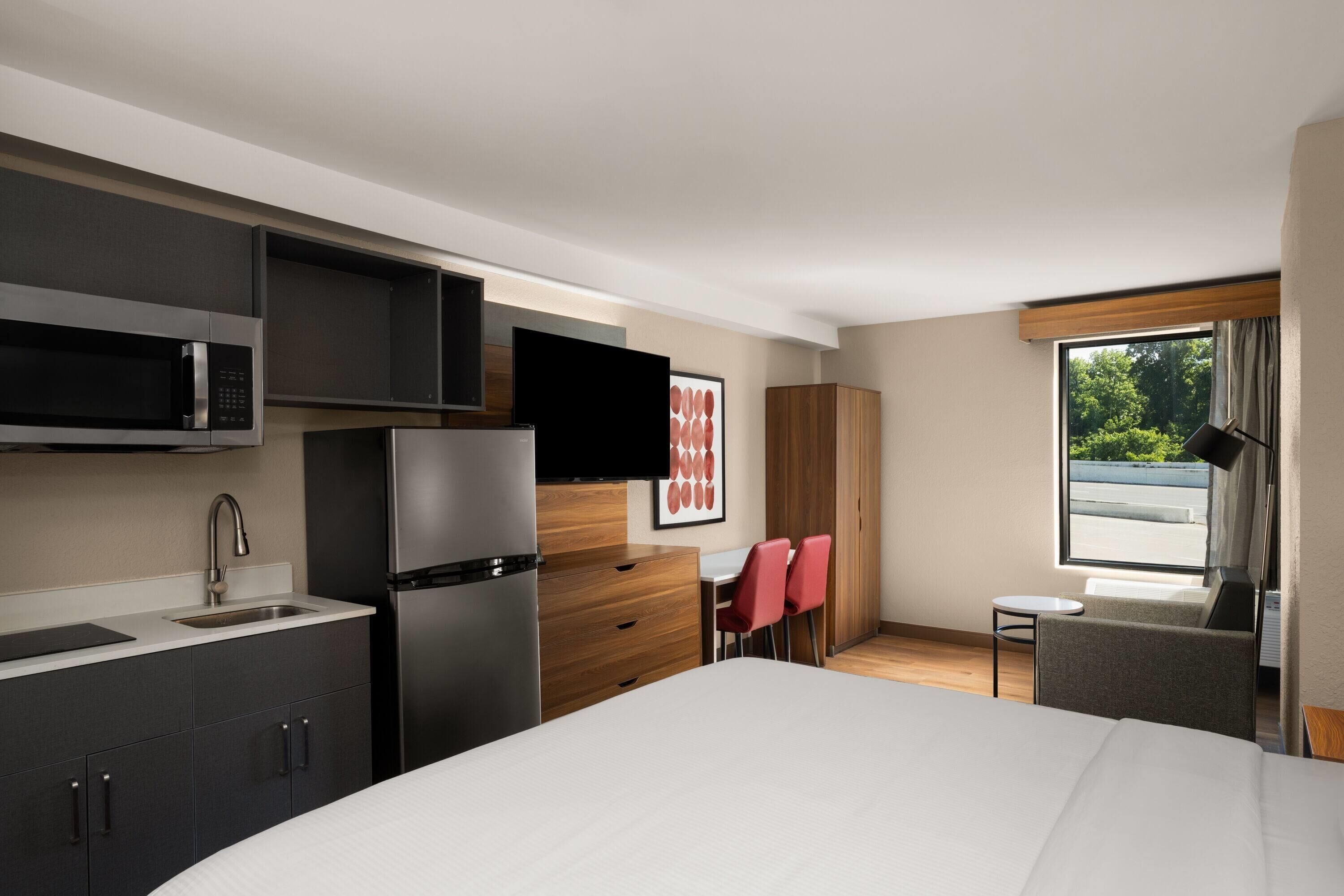 Una habitación de hotel moderna con cocina, cama, pequeña mesa de comedor y ventana con vistas.