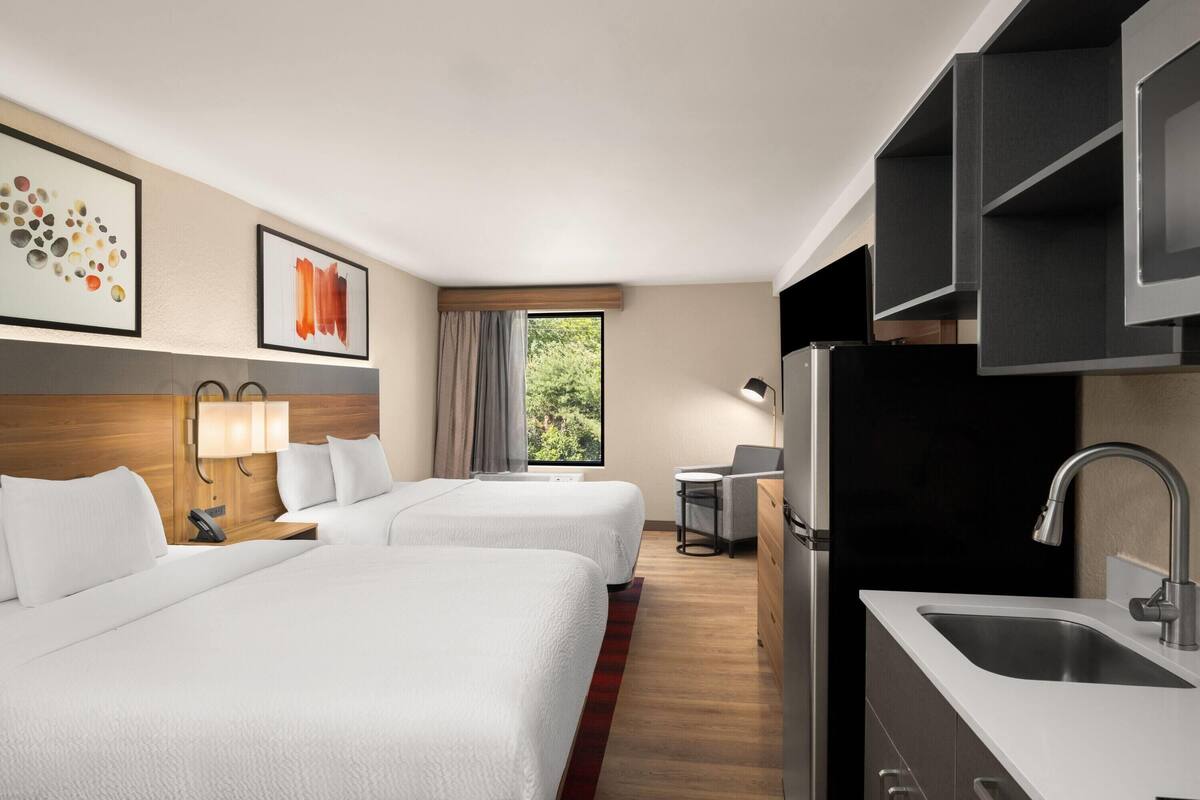 Quarto de hotel com duas camas, cozinha compacta, janela com vista e obras de arte nas paredes.