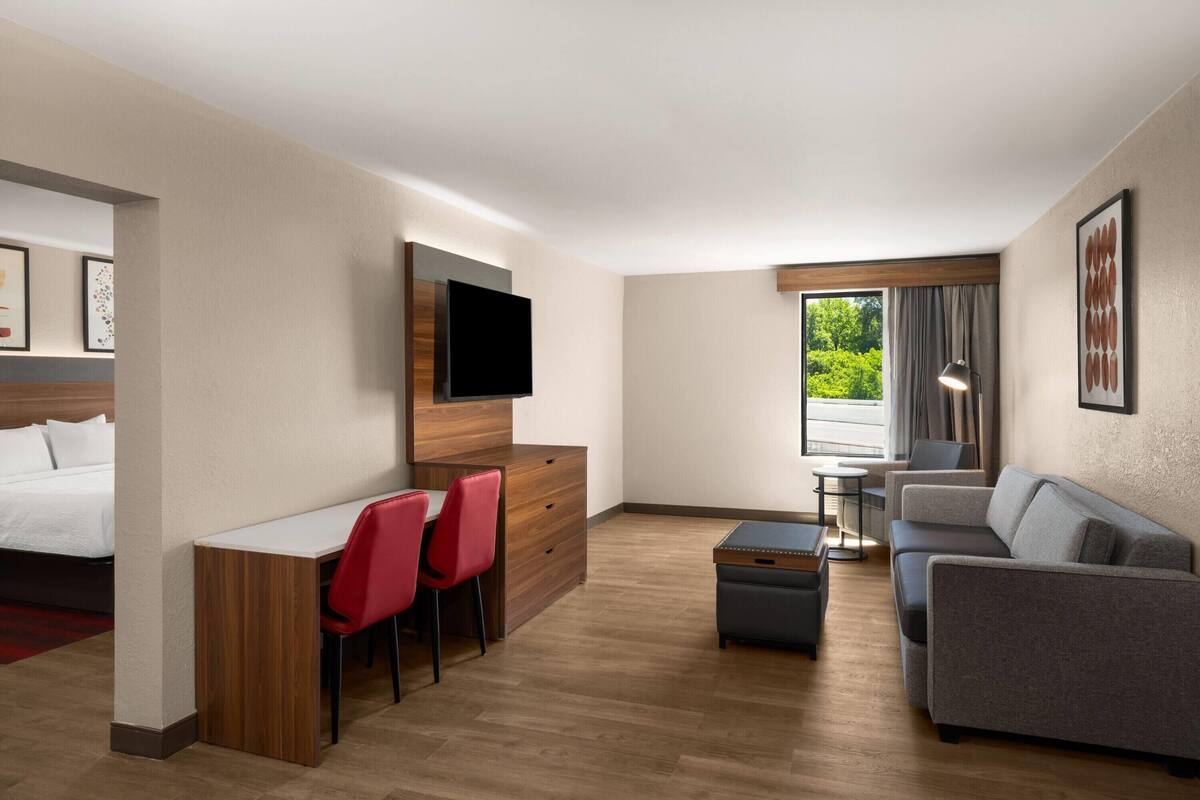 Quarto de hotel com cama, escrivaninha com duas cadeiras vermelhas, sofá cinza, televisão na parede e janela com cortinas.