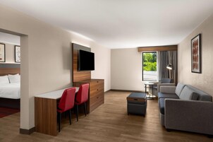 Habitación de hotel con cama, escritorio con dos sillas rojas, sofá gris, televisor montado en la pared y ventana con cortinas.