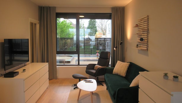 Een moderne woonkamer met een flatscreen televisie, een groene bank, een witte salontafel en een groot raam met uitzicht.