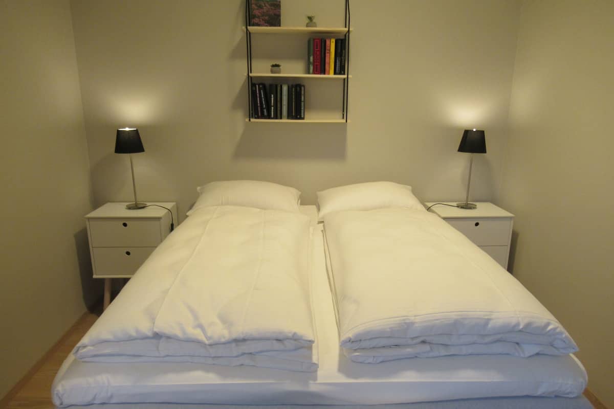 A twin bed with white bedding, two bedside tables with lamps, a small shelf with books, and a towel on the floor.