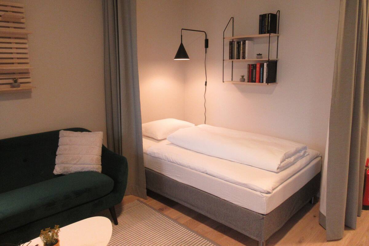 A small, neatly arranged room with a bed, a green sofa, a bedside table, a lamp, and a bookshelf.