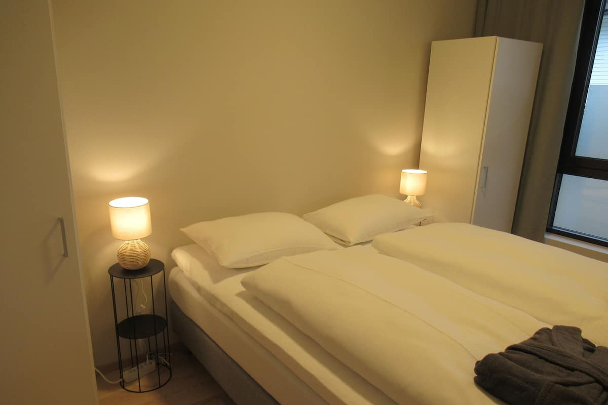 A bedroom with a bed, two bedside lamps, a wardrobe, and a window.