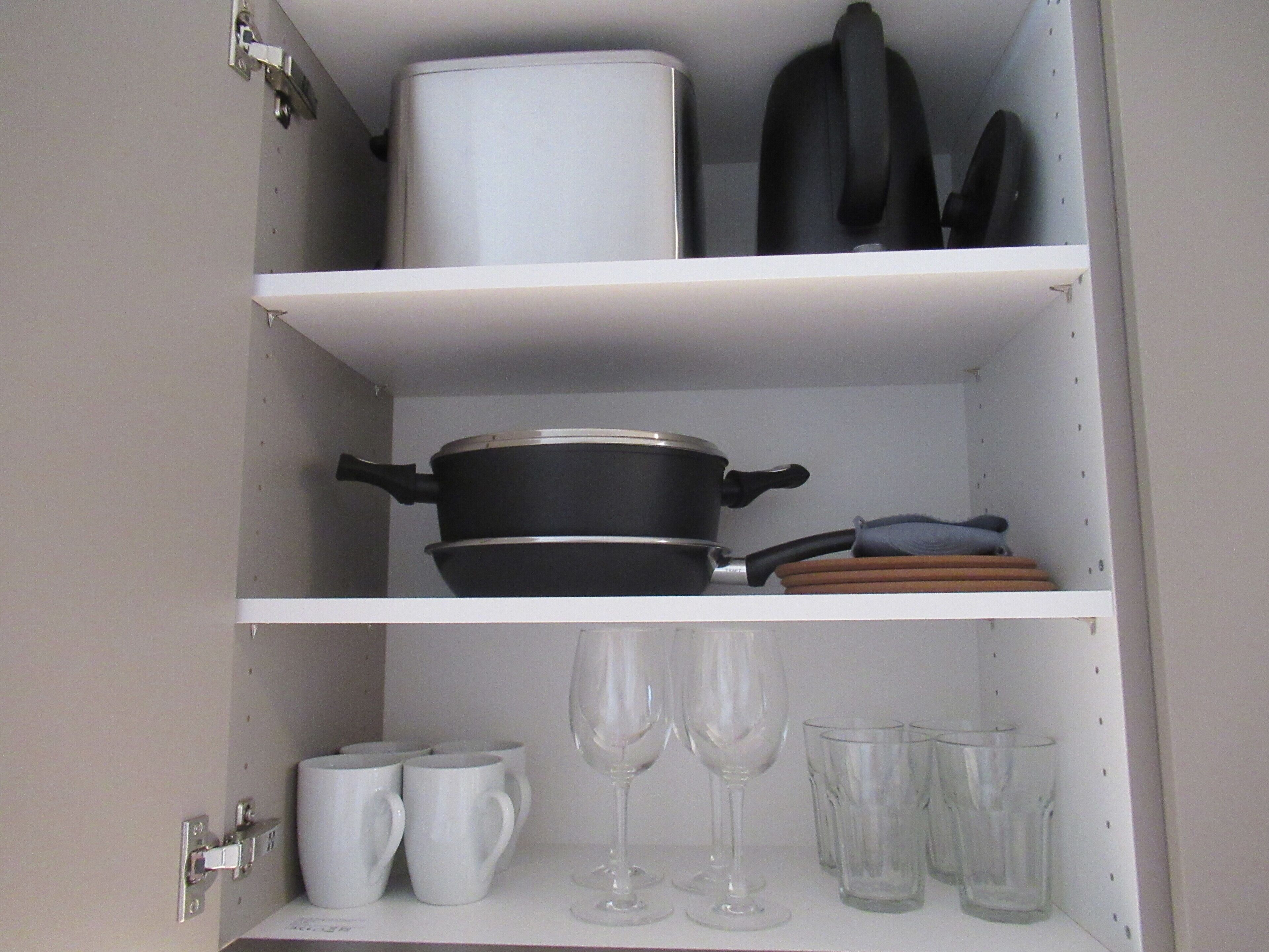 Ett kök med vita hyllor, en svart kastrull, vita muggar och genomskinliga glas.