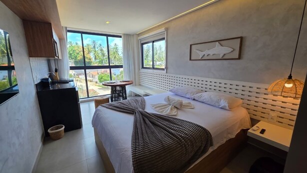 Quarto de hotel moderno com uma cama grande, uma pequena escrivaninha, um banco e vista para palmeiras.