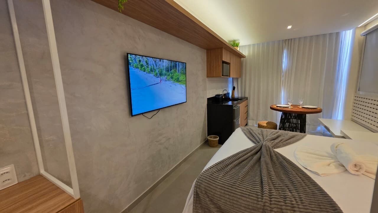 A modern hotel room with a flat-screen TV, a dining table, a kitchen area, and a bed with a blanket.