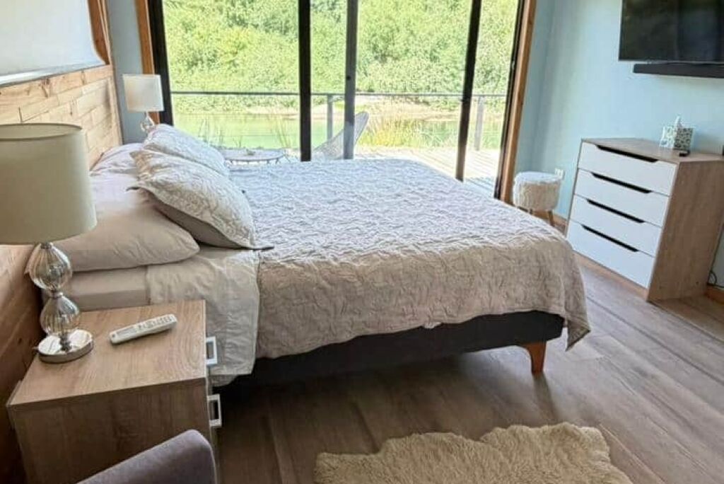 Une chambre à coucher avec un lit, une table de chevet, une lampe et une vue sur la verdure à l’extérieur.