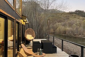 A deck with wicker chairs and tables, overlooking a river and hills.