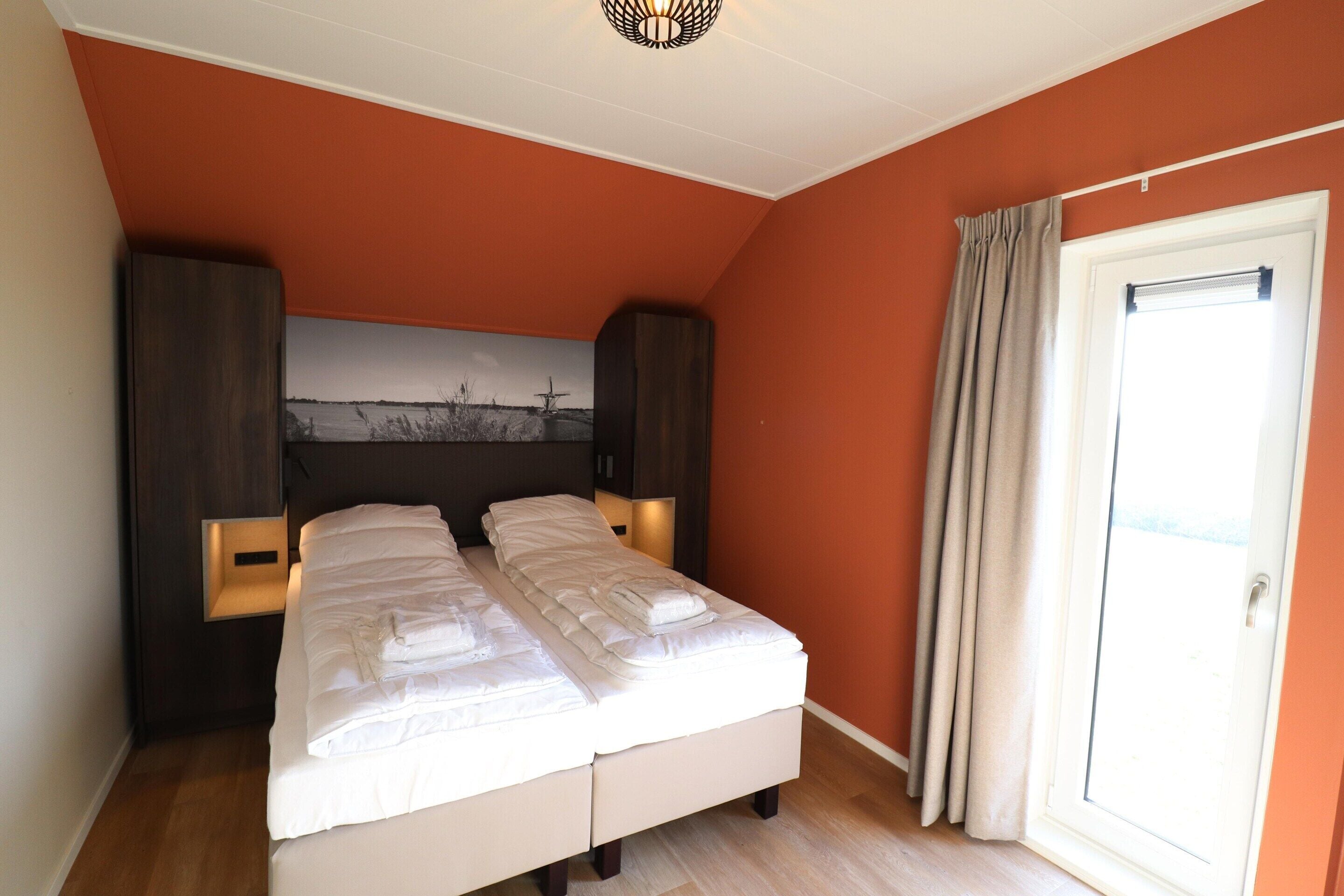 A bedroom with a bed, two bedside tables, a nightstand, a wall-mounted lamp, and a window with curtains.