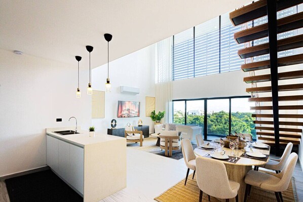 A modern kitchen with a central island, dining area with a round table and chairs, and large windows offering a view of greenery.