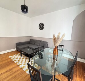 Un salon moderne avec une table à manger en verre, des chaises en bois foncé, un canapé d’angle gris, une petite table basse, une horloge noire et un luminaire suspendu.