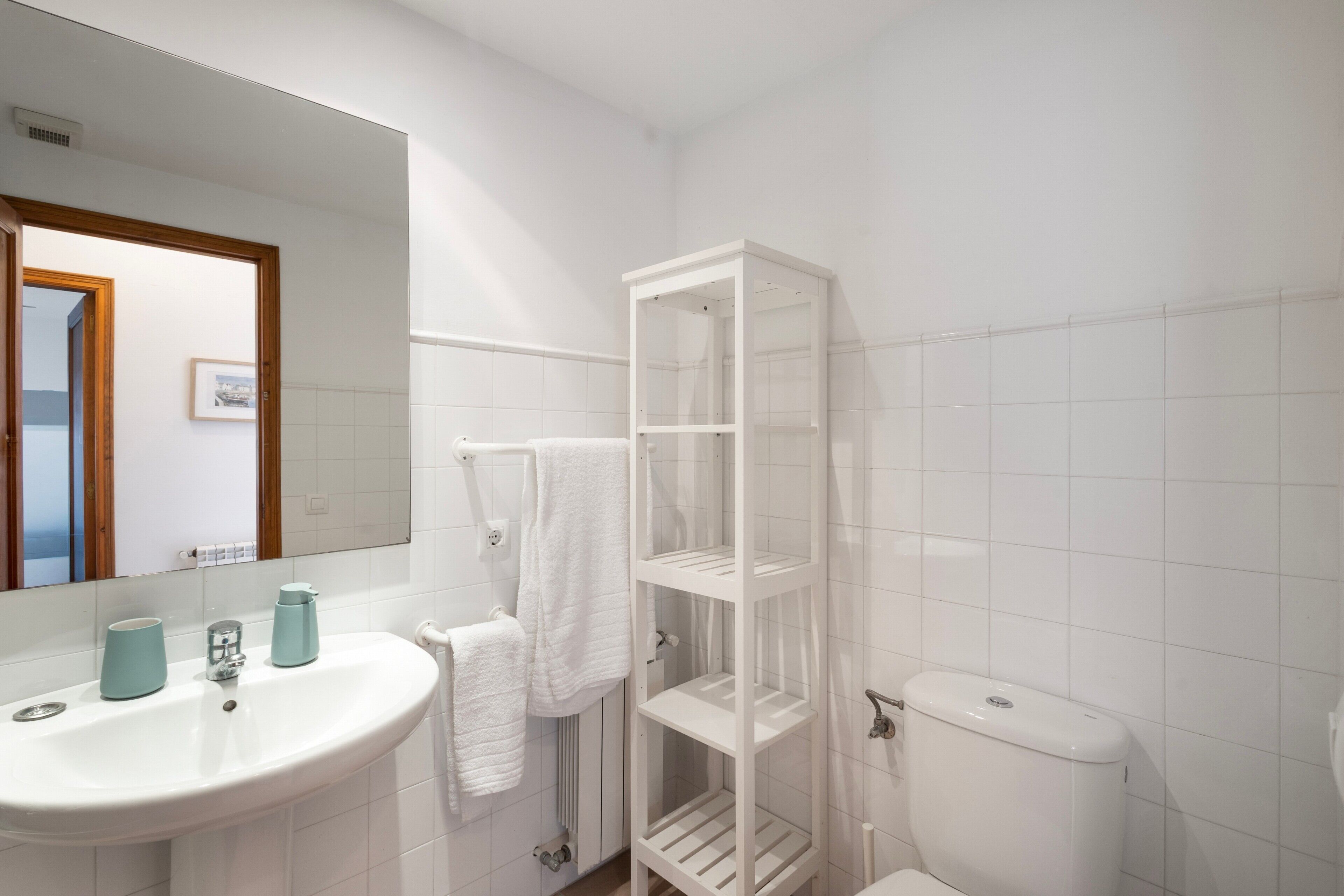 A bathroom with a white sink, mirror, and toilet, and a white shelving unit.