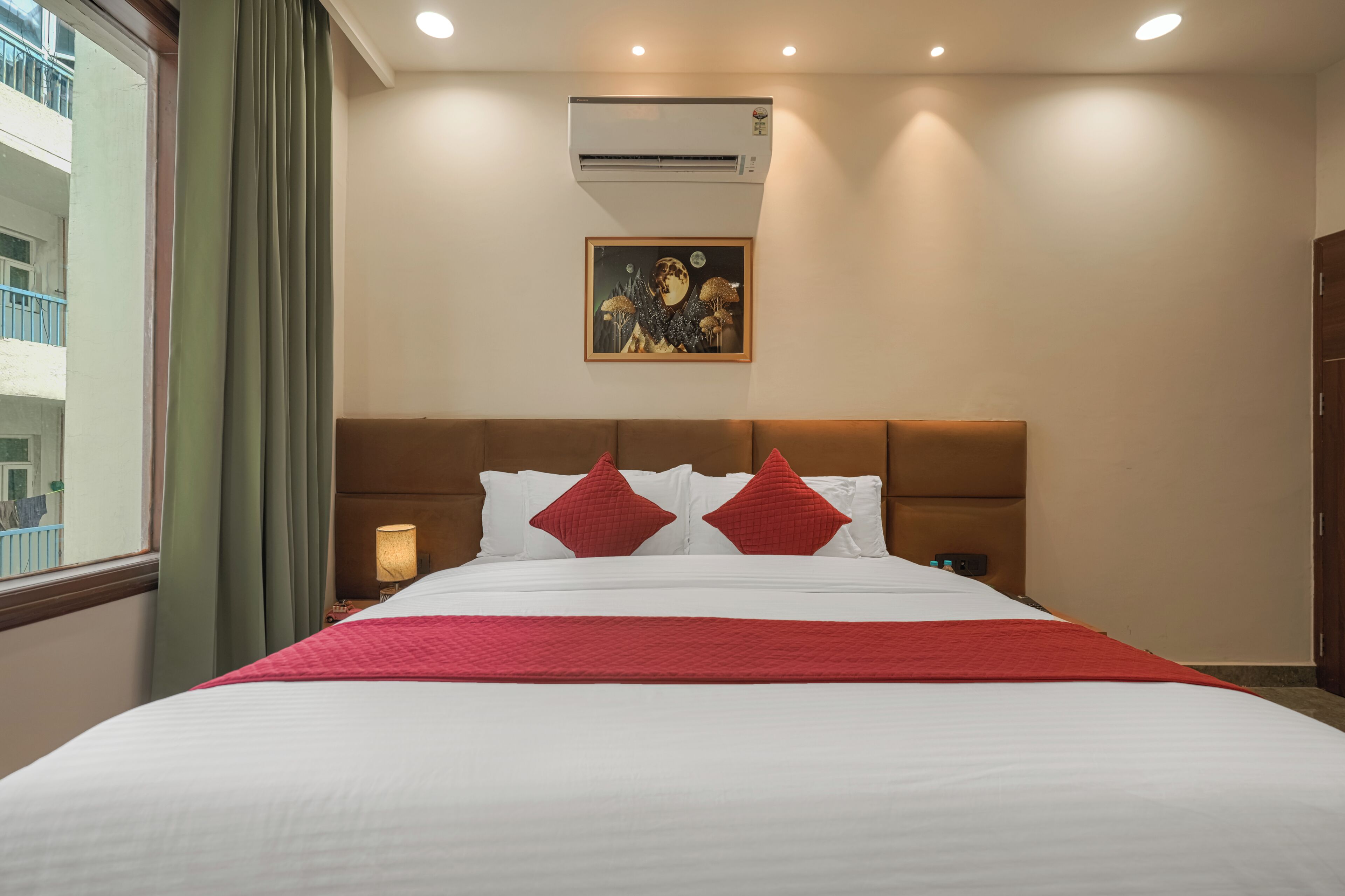 Quarto de hotel com cama, mesa de cabeceira, ar condicionado, um quadro na parede e uma janela com cortinas.