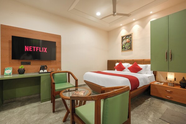 Une chambre d’hôtel avec un lit, un bureau sur lequel est posée une télévision diffusant du contenu « NETFLIX », un fauteuil vert, une petite table et une table de chevet avec une lampe.
