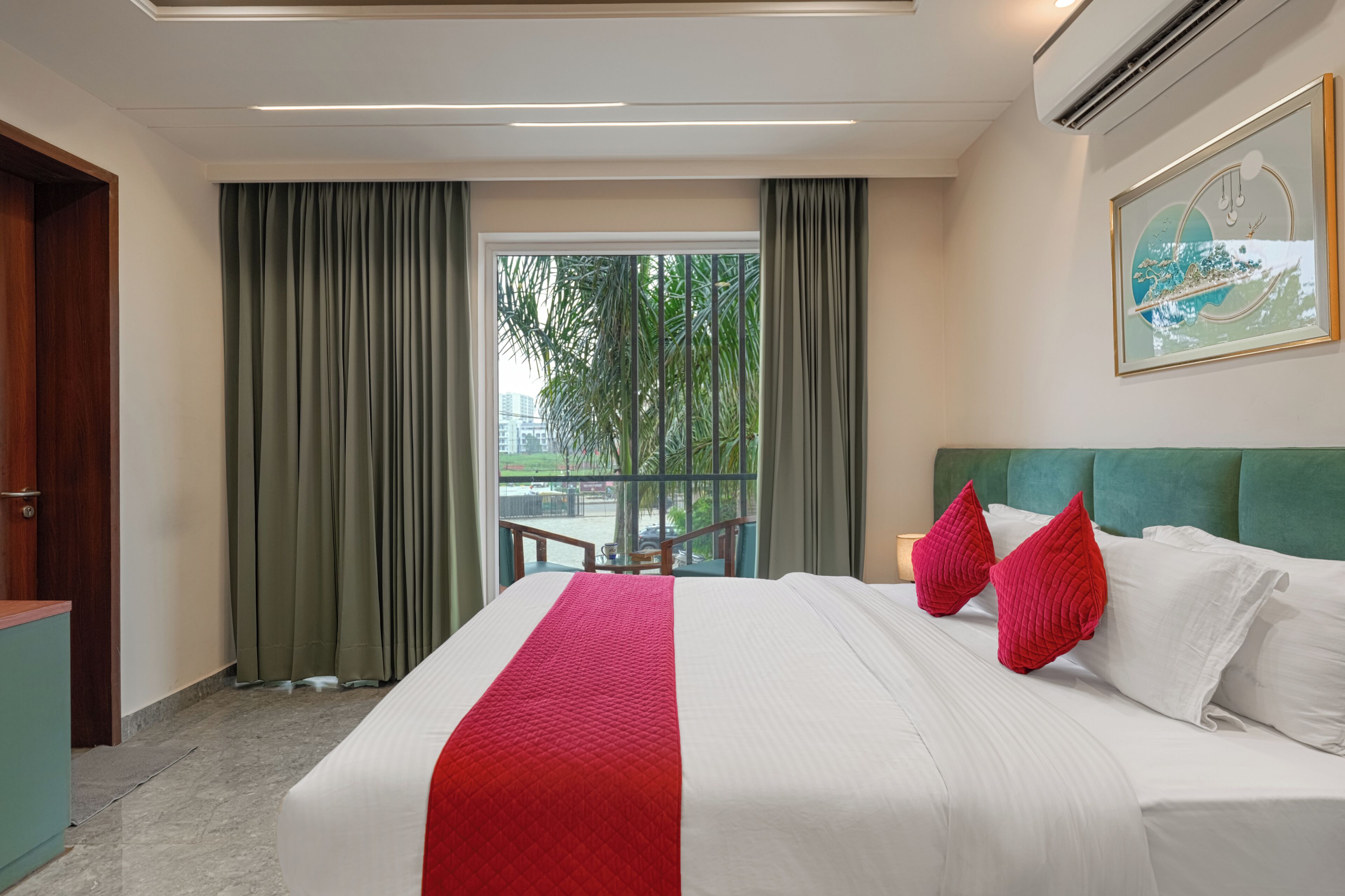 A hotel room with a bed, a view of greenery outside, and a painting on the wall.