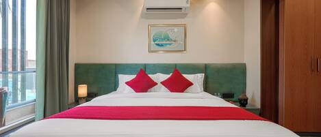 A hotel room with a large bed, a headboard, an air conditioning unit, a framed picture on the wall, and a window with curtains.