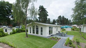 Een gelijkvloers gebouw met een overdekt terras, omgeven door een verzorgd gazon en bomen.