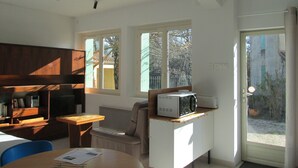 Ett kompakt kök med en mikrovågsugn på en bänkskiva, ett matbord i trä, en stol och ett fönster med utsikt över träd och ett staket.