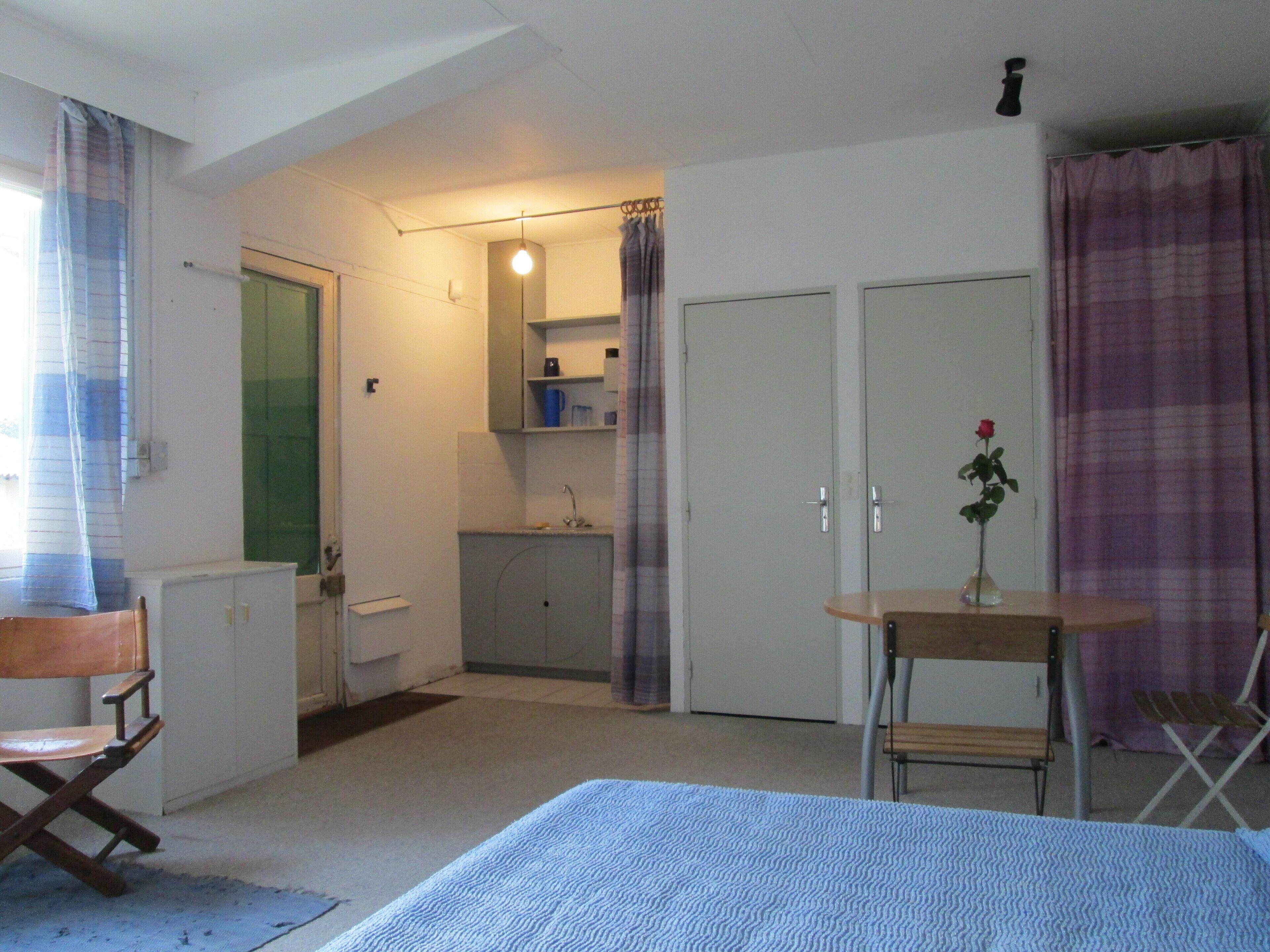 A compact room with a bed, a small table with a vase, a chair, a desk, a wardrobe, a mirror, a sink, and a shower.