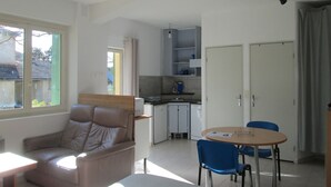 Ett kompakt bostadsutrymme med ett litet kök, en matplats med två stolar, en soffa och ett fönster som släpper in naturligt ljus.