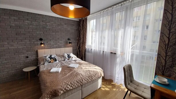 A modern bedroom with a brick wall, a large bed with patterned pillows, a round table with a chair, and a large window with sheer curtains. - Holiday Apartment for 2 People, Swinoujscie (Swinoujscie)